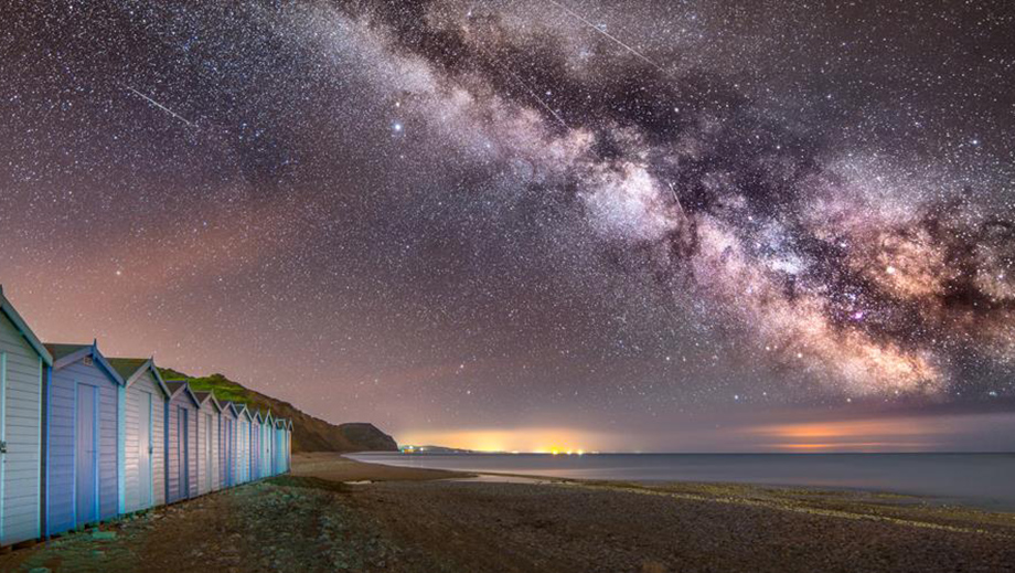 A long exposure night-time photograph of the Milky Way over Charmouth Beach in Dorset, UK. The horizon on the right is lit by lights on a distant stretch of coast, and on the left is a row of beach huts in pastel blues.