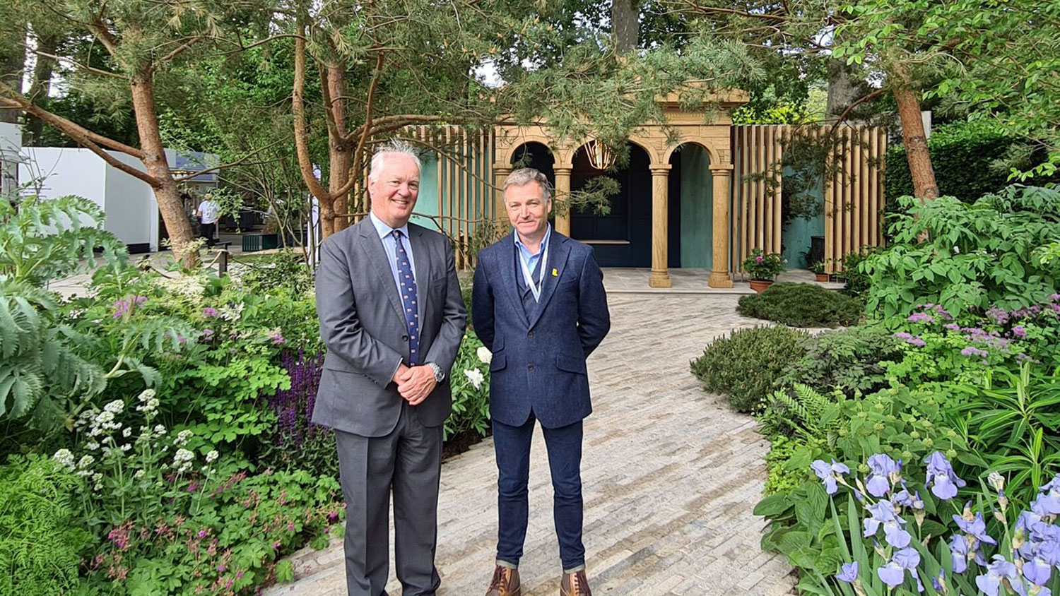 Garden Designer Chris Beardshaw and RNLI Chief Executive Mark Dowie dressed in suits, standing on the Purbeck stone paving in the RNLI Garden at the 2022 RHS Chelsea Flower Show. Design features include the Georgian pavilion and its beacon in the background and the calming green, blue and purple hues of the flowers and plants in the foreground.  