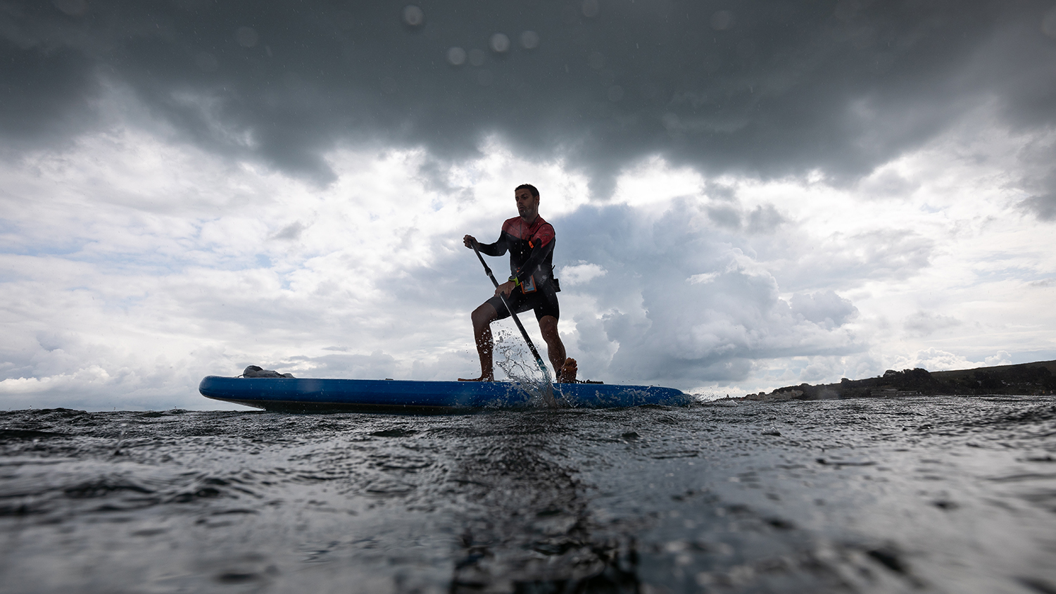 A paddleboarder standing on his blue board, surrounded by grey sky and sea