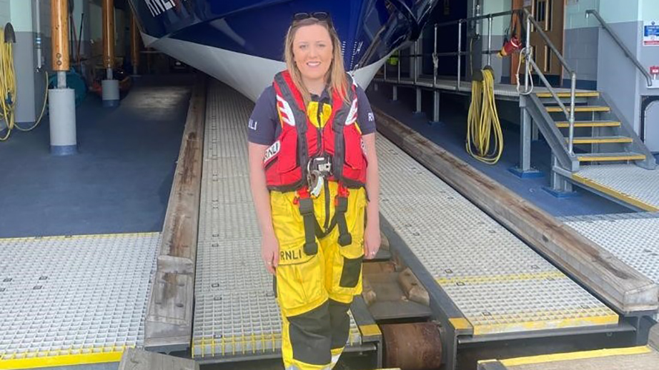 Volunteer Lifeboat Crew Member Caryl Jones in crew kit, smiling in front of the Porthdinllaen Lifeboat Station, with the shiny Tamar lifeboat in the background
