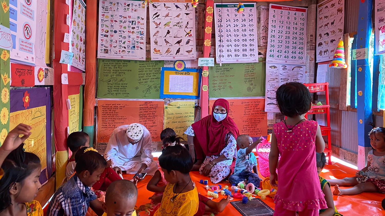 Inside a Barisal Anchal, a small room decorated with bright walls and posters, with several children sitting inside 