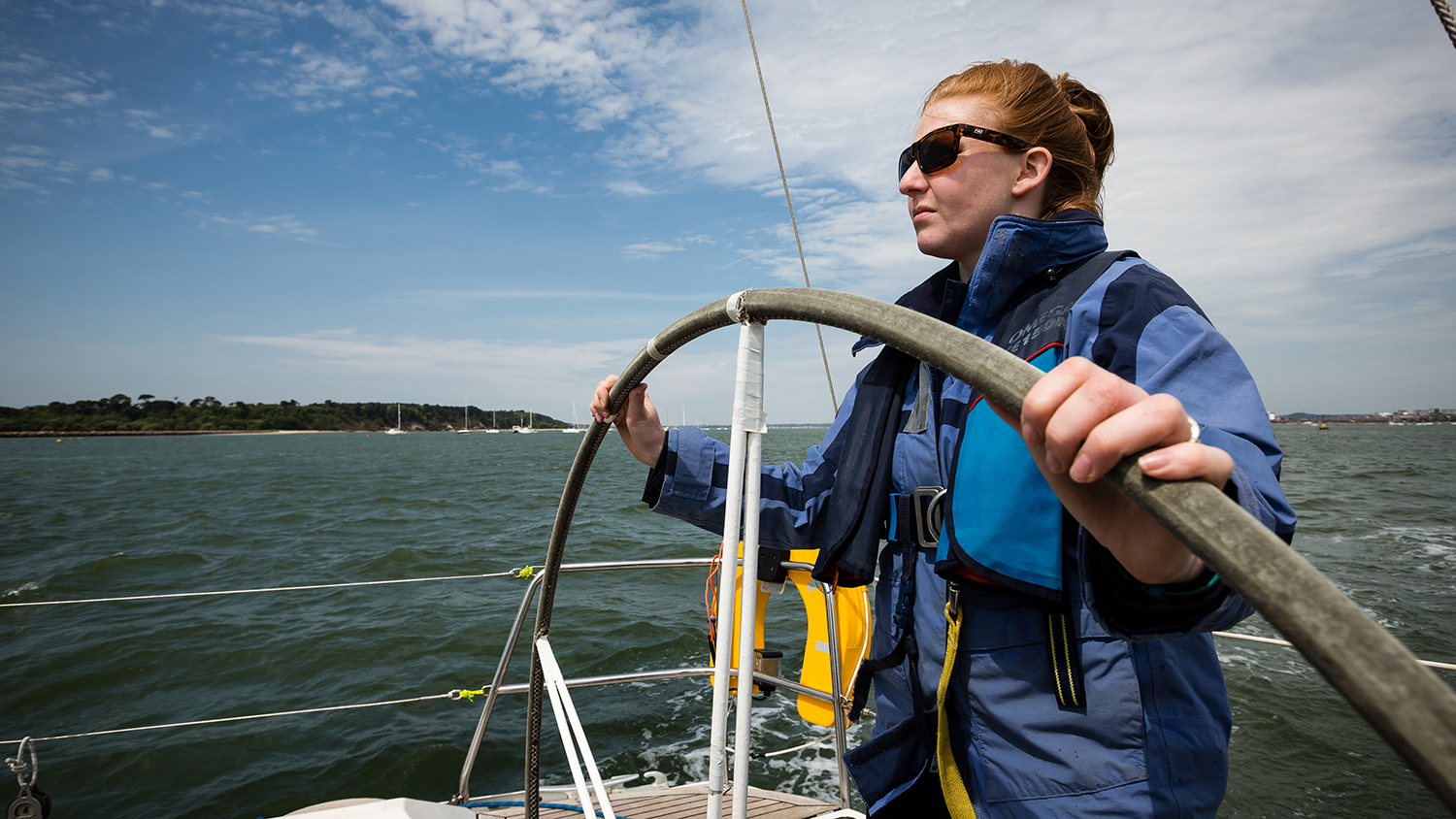 A woman steering a yacht, wearing sunglasses and a blue jacket