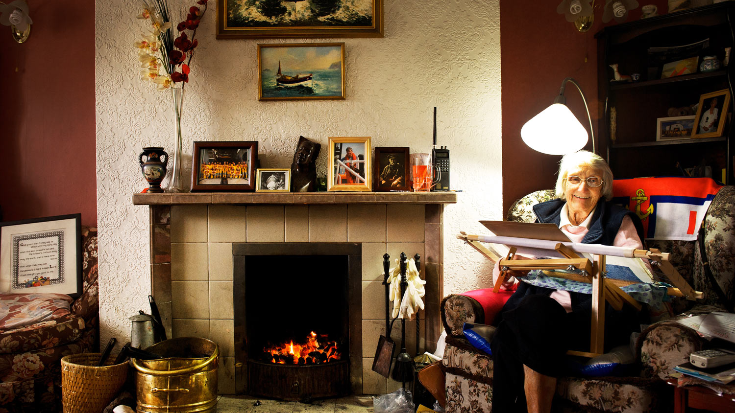 An elderly woman sits knitting items next to a fireplace