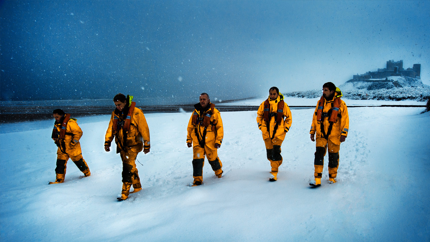 Five lifeboat crew members walk across a snowy beach with a castle in the background