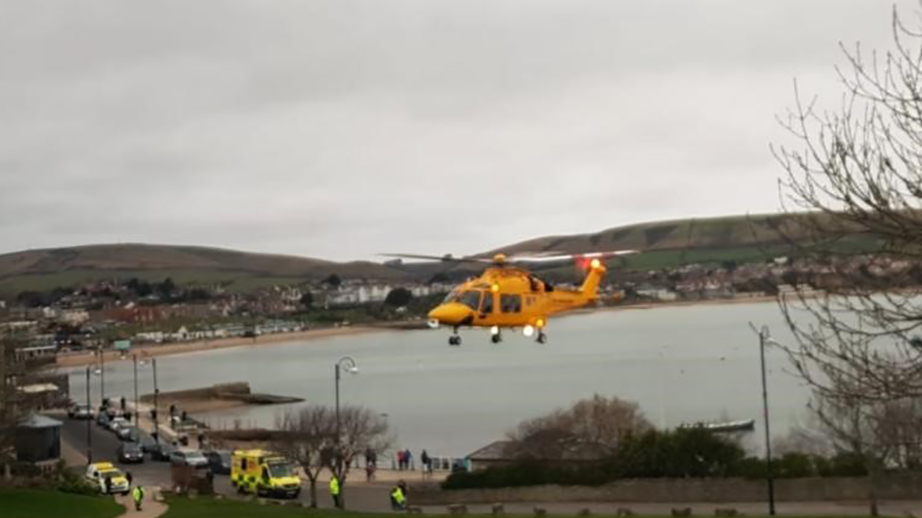 A yellow air ambulance flies over Swanage. You can see an ambulance and several people wearing high-vis jackets below.