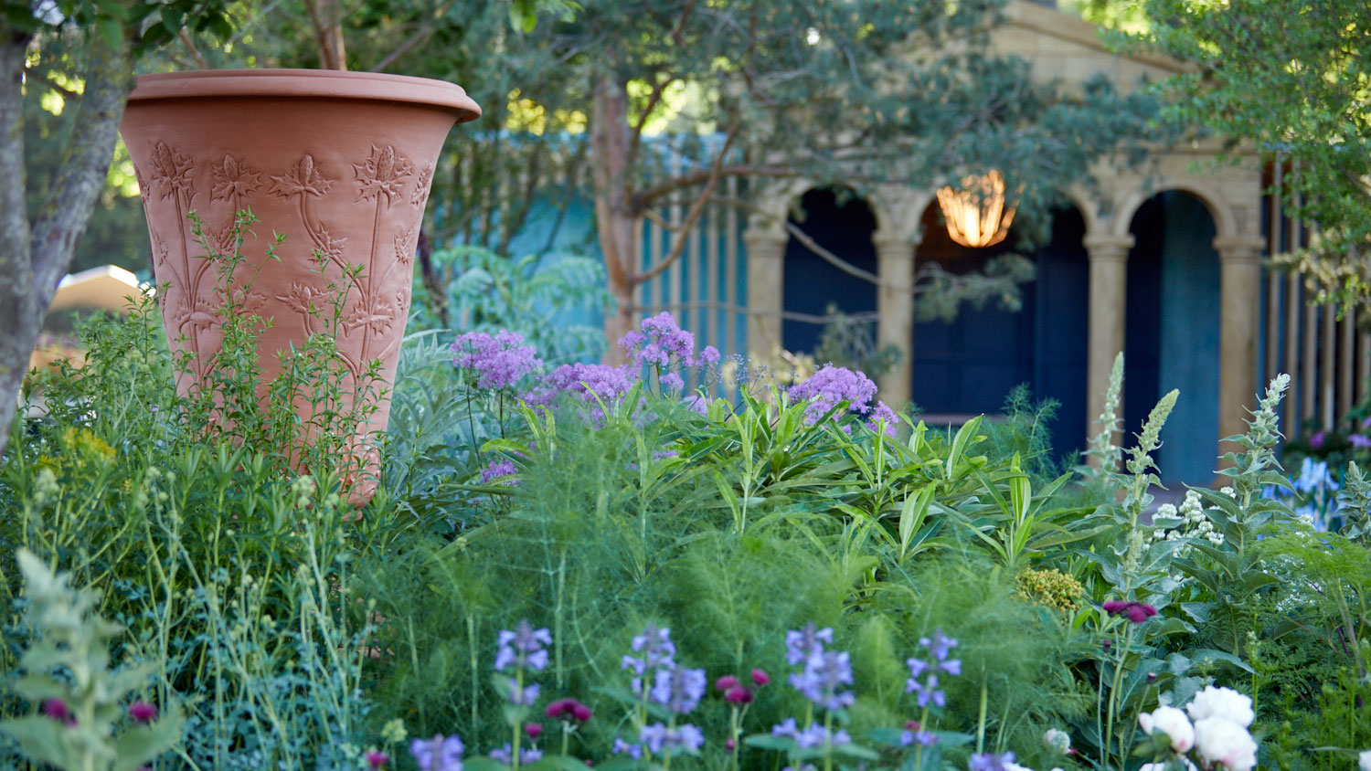 The RNLI Garden at the 2022 RHS Chelsea Flower Show. Photo shows the Georgian pavilion and its beacon in the background, and the calming green, blue and purple hues of the flowers and plants, with a terracotta urn in the foreground.  