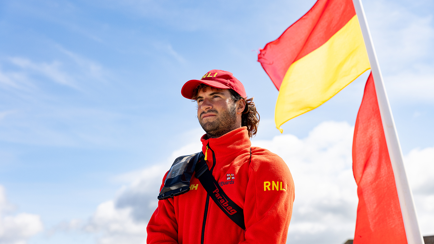 Senior lifeguard Nick Ellison in red RNLI fleece top and cap standing next to red and yellow flag, looking out to sea