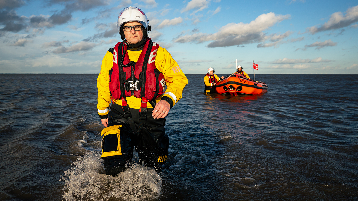 Crew Members Phil and Matt stand knee-deep in the water, with Steve at the helm in the lifeboat
