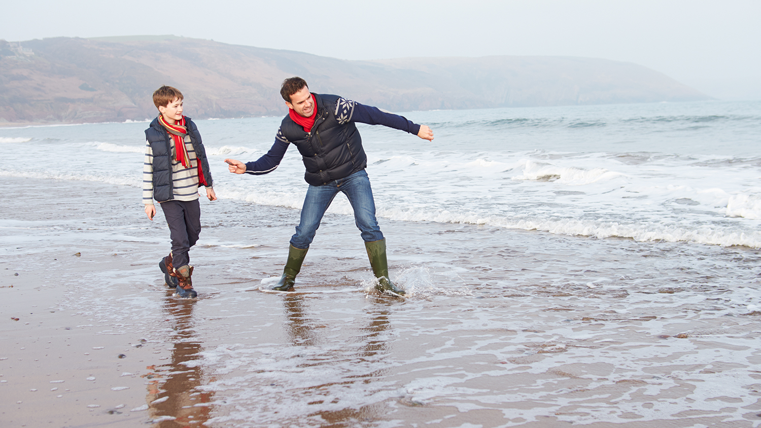A man and a boy, wrapped up in jackets and wellies, skim stones on the beach