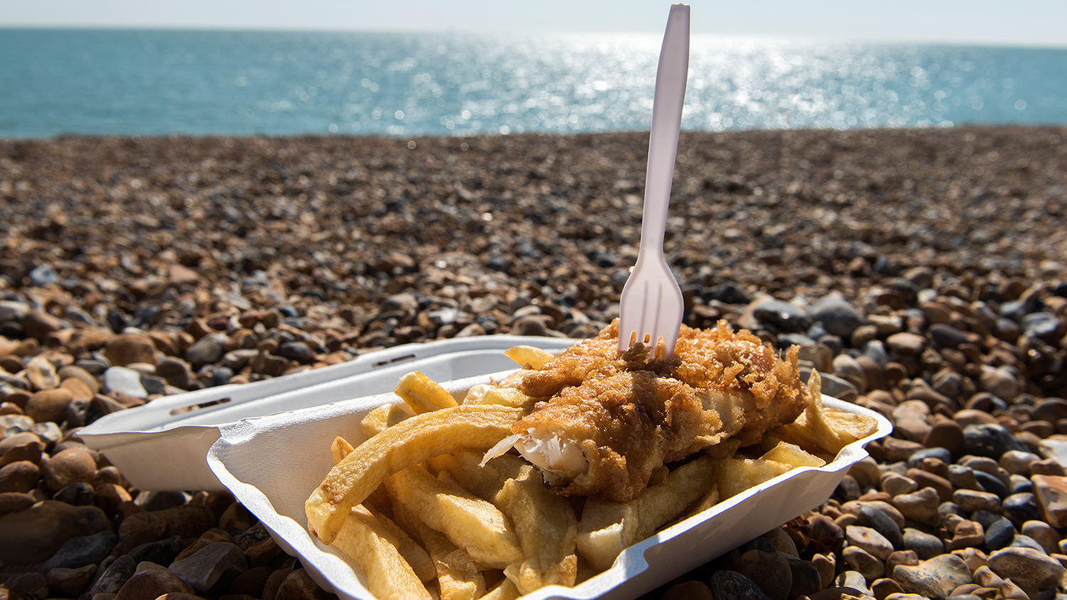 A white, polystyrene container of fish and chips, resting on a pebbled beach