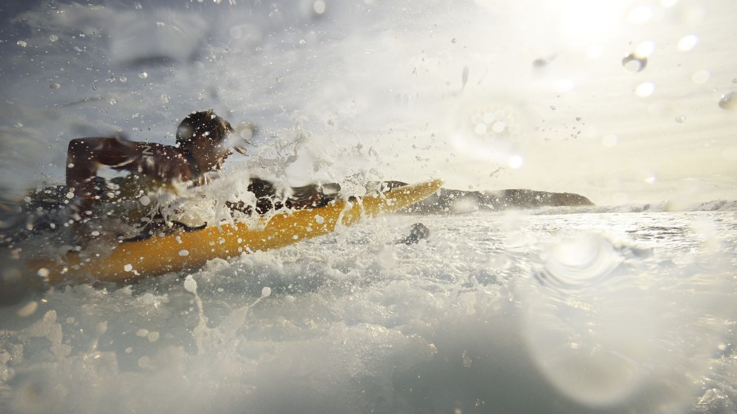 An RNLI lifeguard paddles out to sea through surf on a rescue board. Water splashes are on the camera.