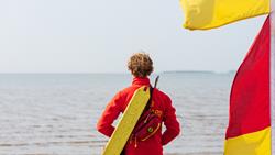 The back of an RNLI lifeguard, looking out to sea. He is wearing a red fleece with a yellow rescue tube over his shoulder and is standing next to a red and yellow flag.