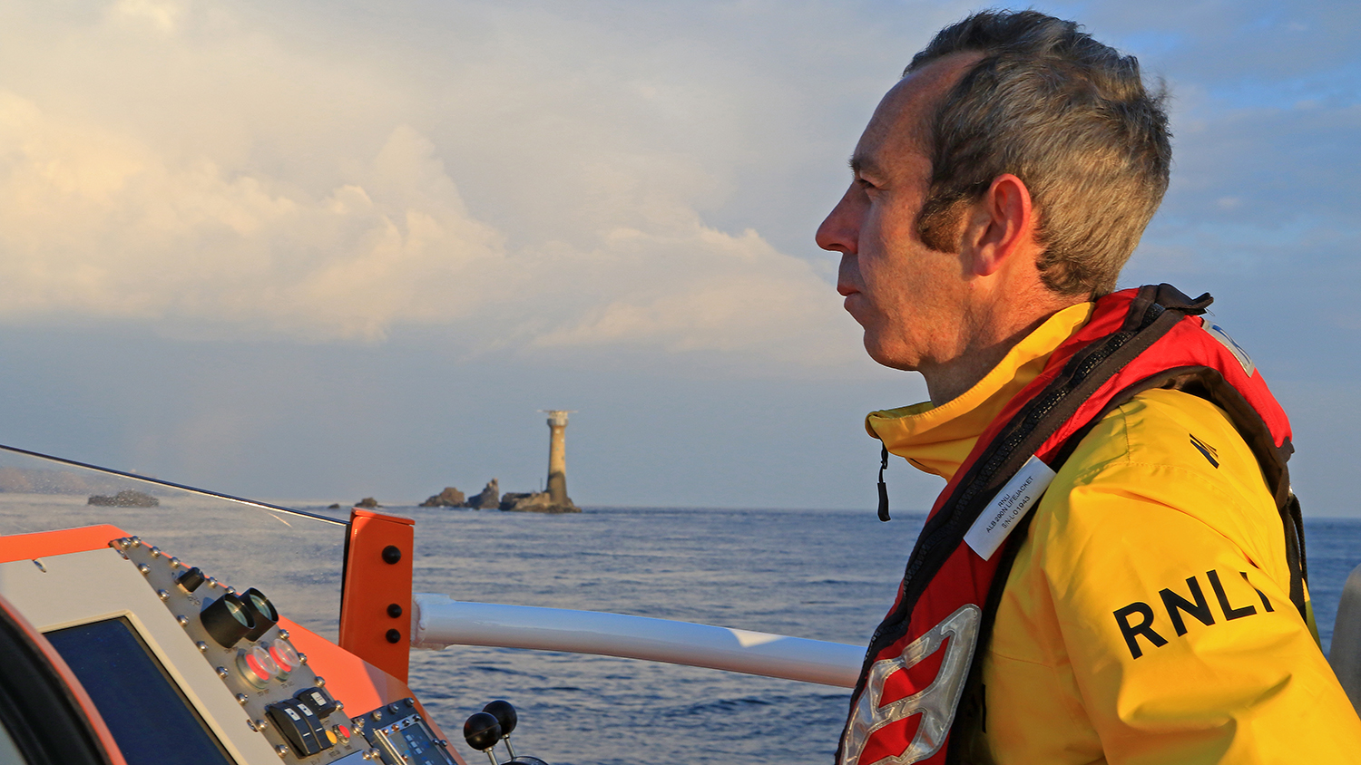 Coxswain Ollie George in profile in lifeboat kit at the helm of the Tamar class lifeboat, with Longships Lighthouse in the background
