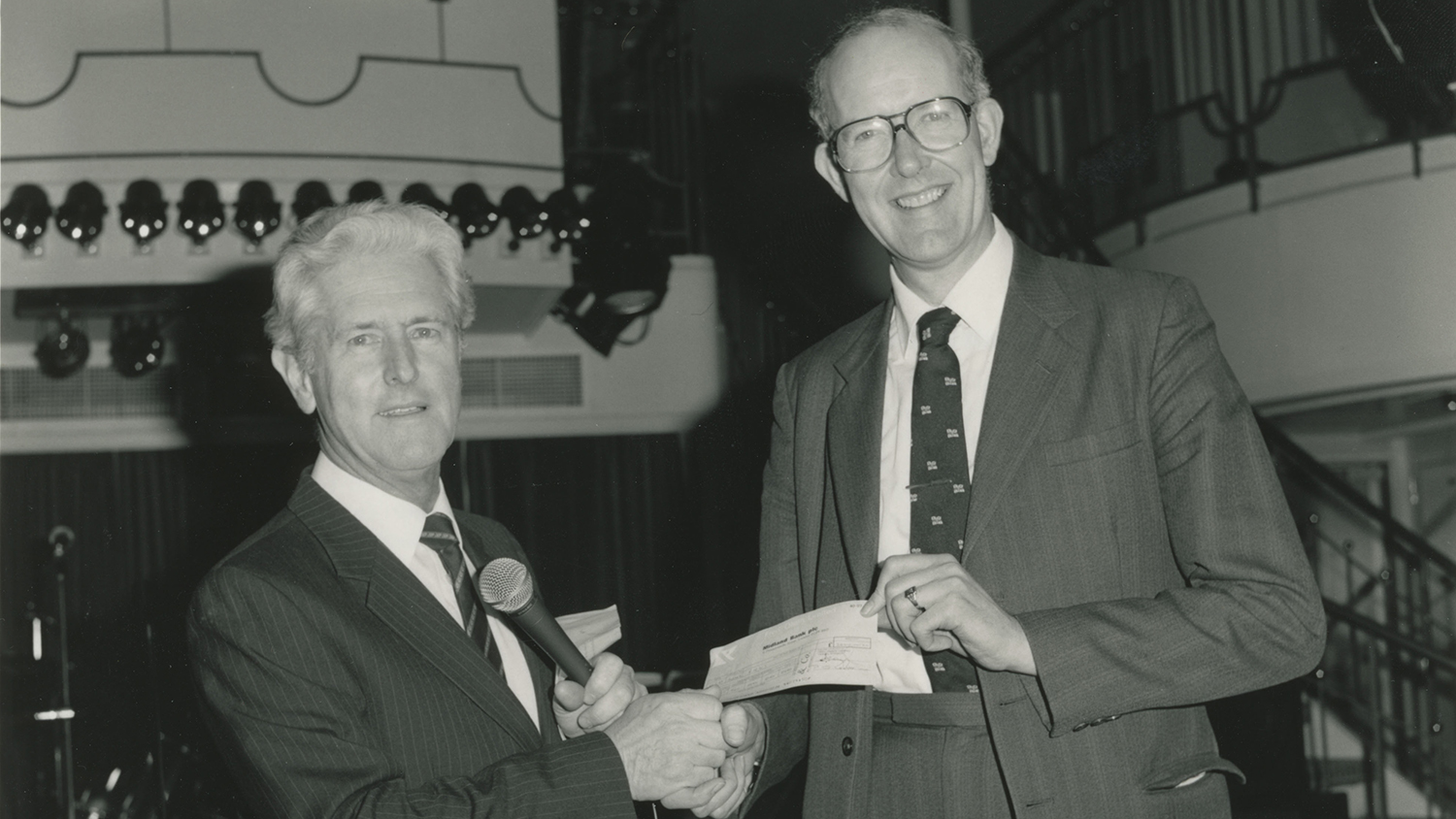 A black and white photograph of two men wearing suits and shaking hands. One of the men is handing a cheque to the other. 