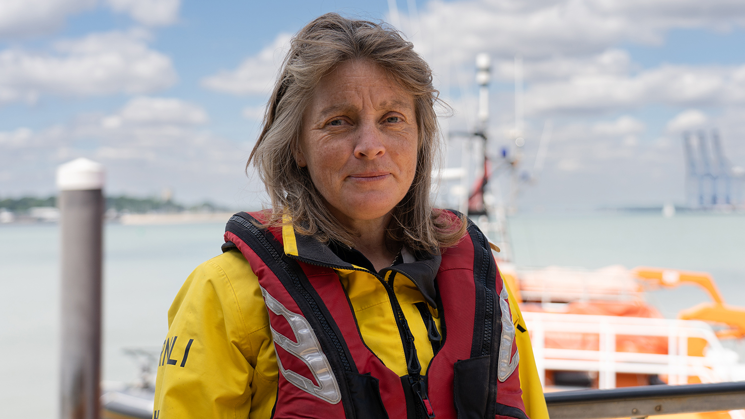A close-up, head-and-shoulders photo of Harwich RNLI Full-time Coxswain Di Bush standing outdoors with part of the moored all-weather lifeboat in the background. Di has long, light brown hair and is wearing her yellow RNLI crew kit including red lifejacket.