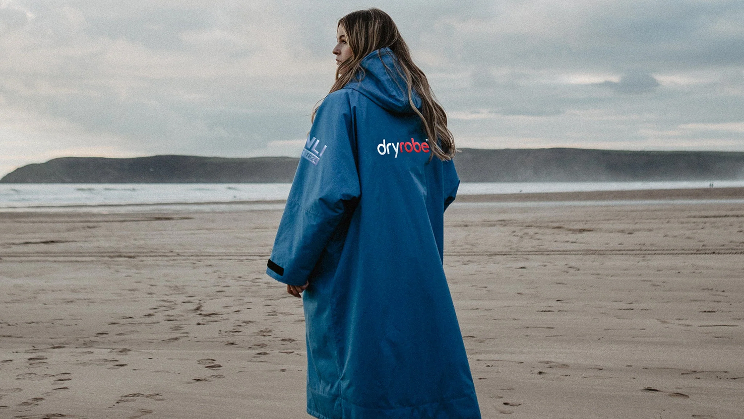 A woman stands on the beach wearing a cosy-looking blue, long-sleeved, hooded changing robe