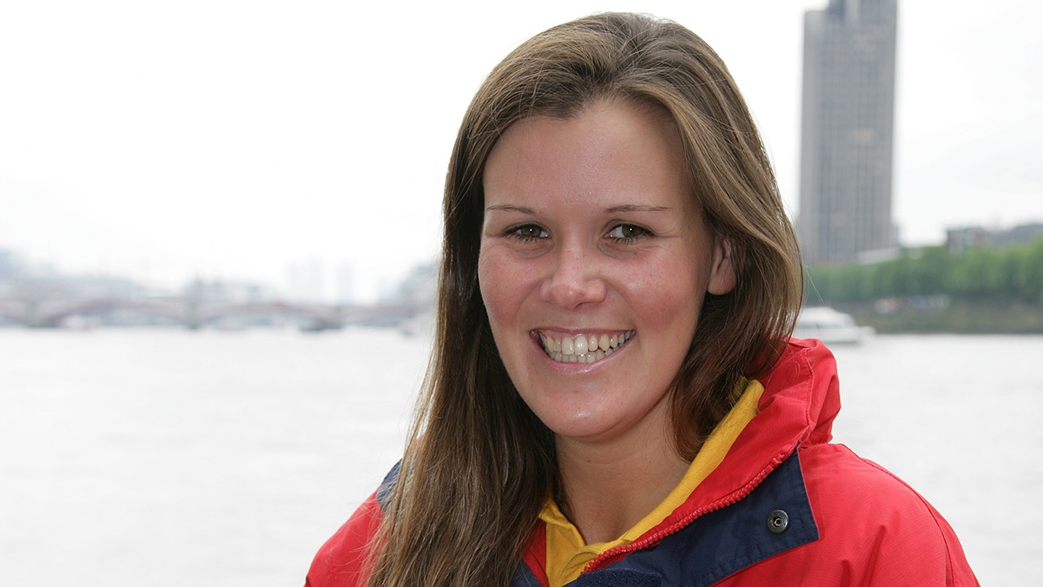 RNLI Lifeguard Sophie Grant-Crookston standing outdoors by a river. Sophie is wearing her RNLI Bronze Medal for Gallantry on her red RNLI lifeguards jacket. She has long brown hair and a beautiful smile. 