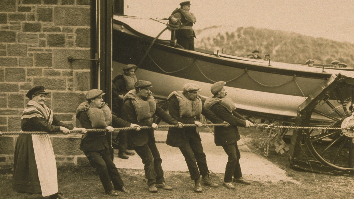 A sepia photo of the launch of Cresswell lifeboat. Margaret Armstrong and four male crew members are holding the rope to slowly release the lifeboat from the boathouse. The coxswain onboard the lifeboat looks ahead.