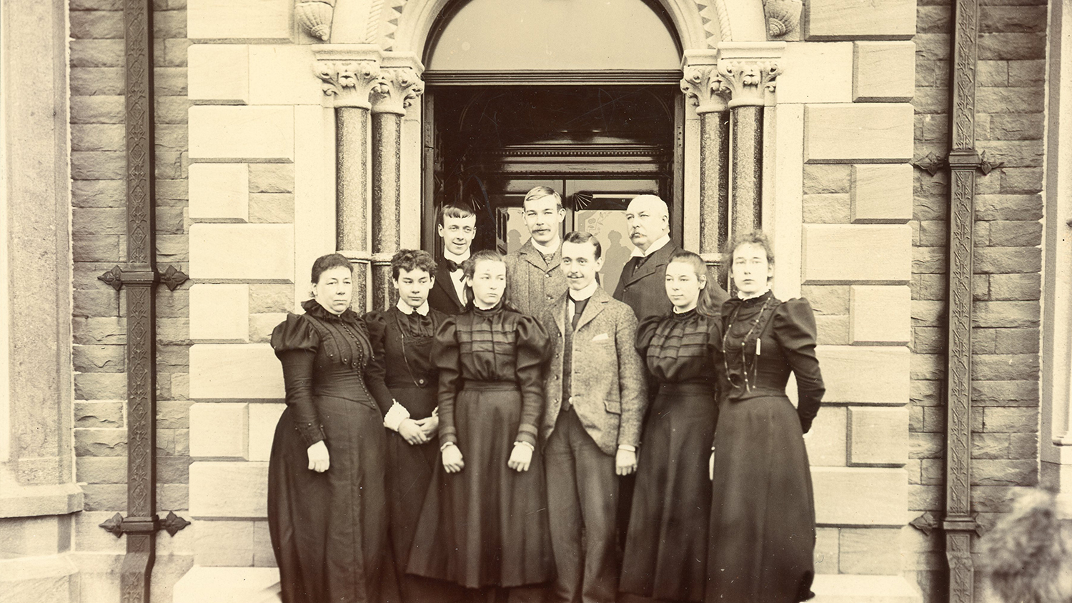 A group sepia photo of the Macara family. Lady Marion Macara and the young female family members are all dressed in long, dark dresses. Sir Charles Macara and the young male family members are dressed in suits. They are standing outside the grand entrance of a building.