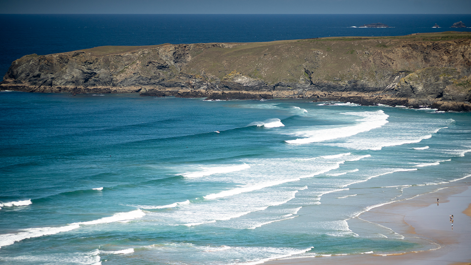 A Cornish bay edged by a rocky headland, with a rip current visible in the centre of the beach by the shoreline – an area of darker and deeper water where the waves are not breaking.