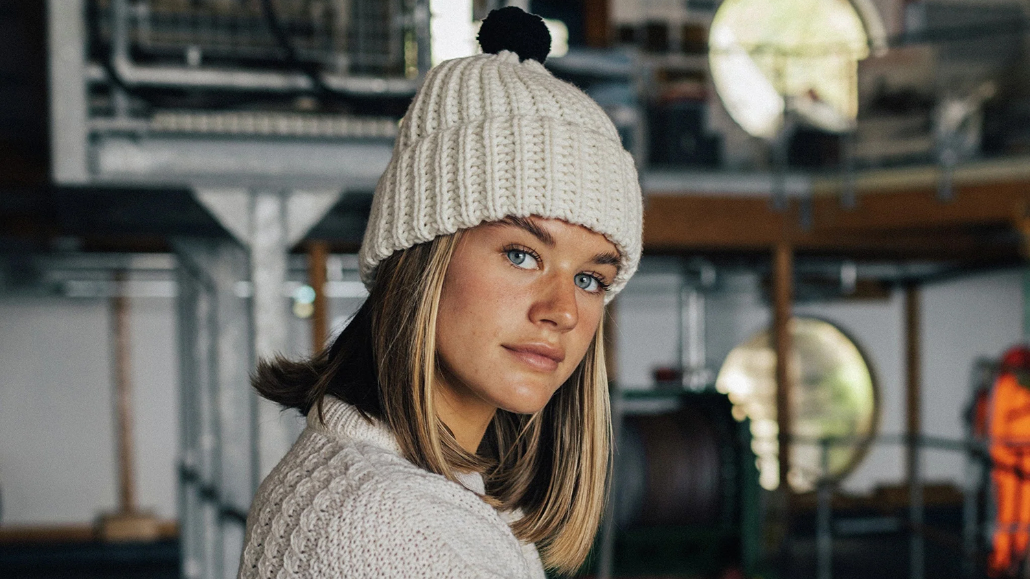 A woman with blonde hair looks at the camera, wearing a cream-coloured beanie with a navy bobble on top