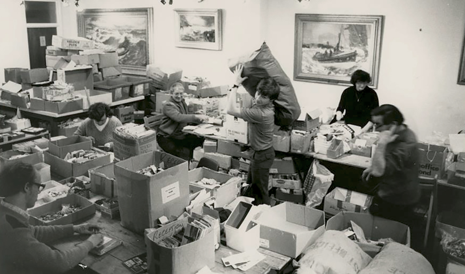 People working at desks sorting through buttons and postcards that were sent in to raise money for the RNLI as part of a Blue Peter appeal. The room is cluttered, with overflowing boxes and sacks, as well as large piles of paperwork.
