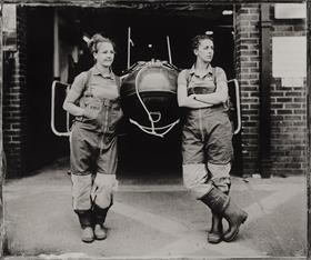 A black and white photograph of two female crew members leaning against the front of an inshore lifeboat, which peeks out from a brick boathouse. They’re both wearing their crew kit, which includes bibbed trousers and wellies.