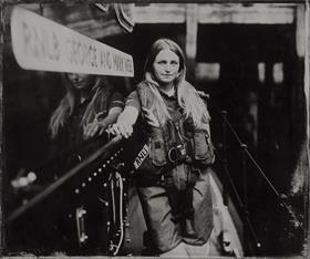 An old-fashioned-looking black and white photograph of Leah Hunter, a volunteer crew member at Whitby RNLI. She’s standing in front of the station’s lifeboat, in her crew kit, looking into the camera and smiling.