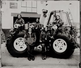 A black and white photograph of five female RNLI crew members. Three of them are sitting on a tractor, while the other two are in front and leaning against it. They’re wearing their crew kit and staring into the camera.