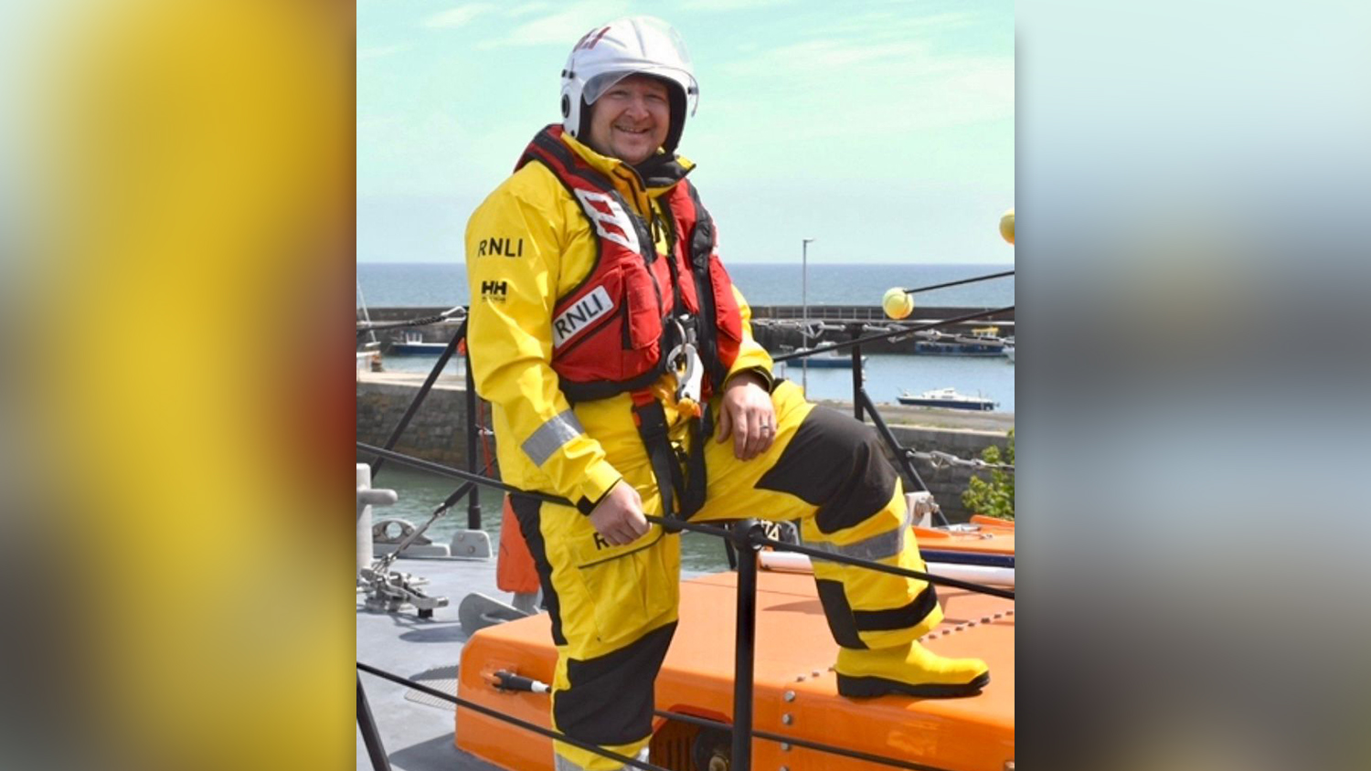 Brendan Rooney, Crew Member at Newcastle RNLI, standing on an all-weather lifeboat in full crew kit. The sky is blue with a few small clouds, and the sea is in the background.