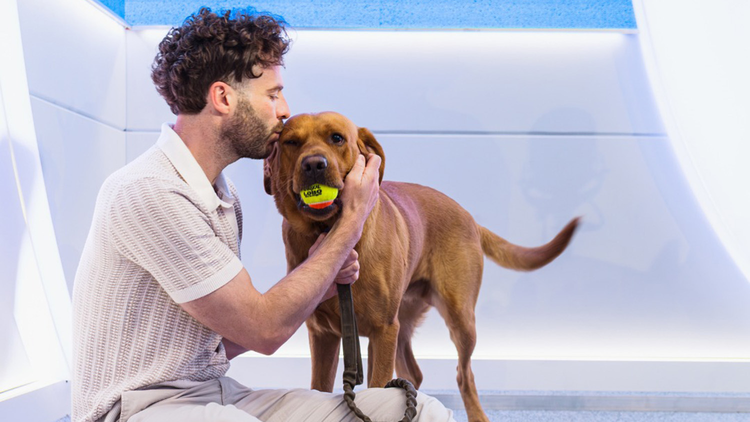 Radio Presenter Jordan North is sat on the floor of a white studio kissing Creed, a red Labrador, who is holding a tennis ball and wagging his tail.