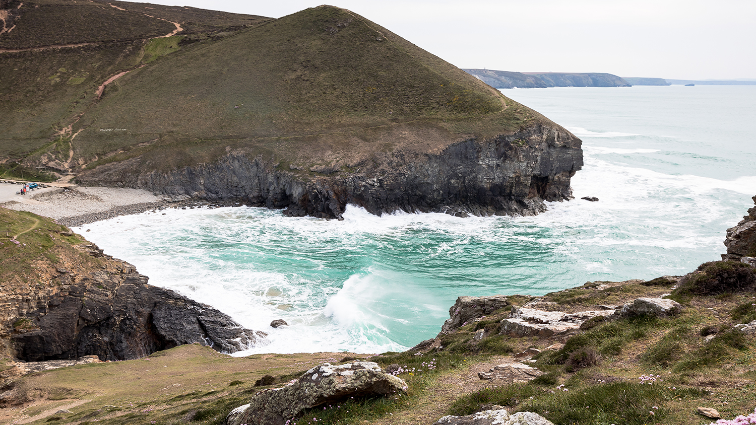 A view from the northern cliffs of Chapel Porth with the beach below. It’s high tide and the churning sea is crashing into the rocks. 