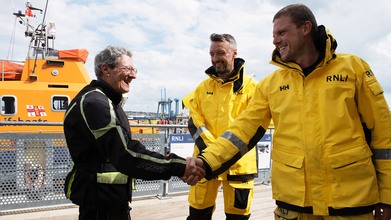 Three men are stood in front of a lifeboat, the man on the left, Dermot, is shaking hands with one of the lifeboat crew members on the right, Chris.