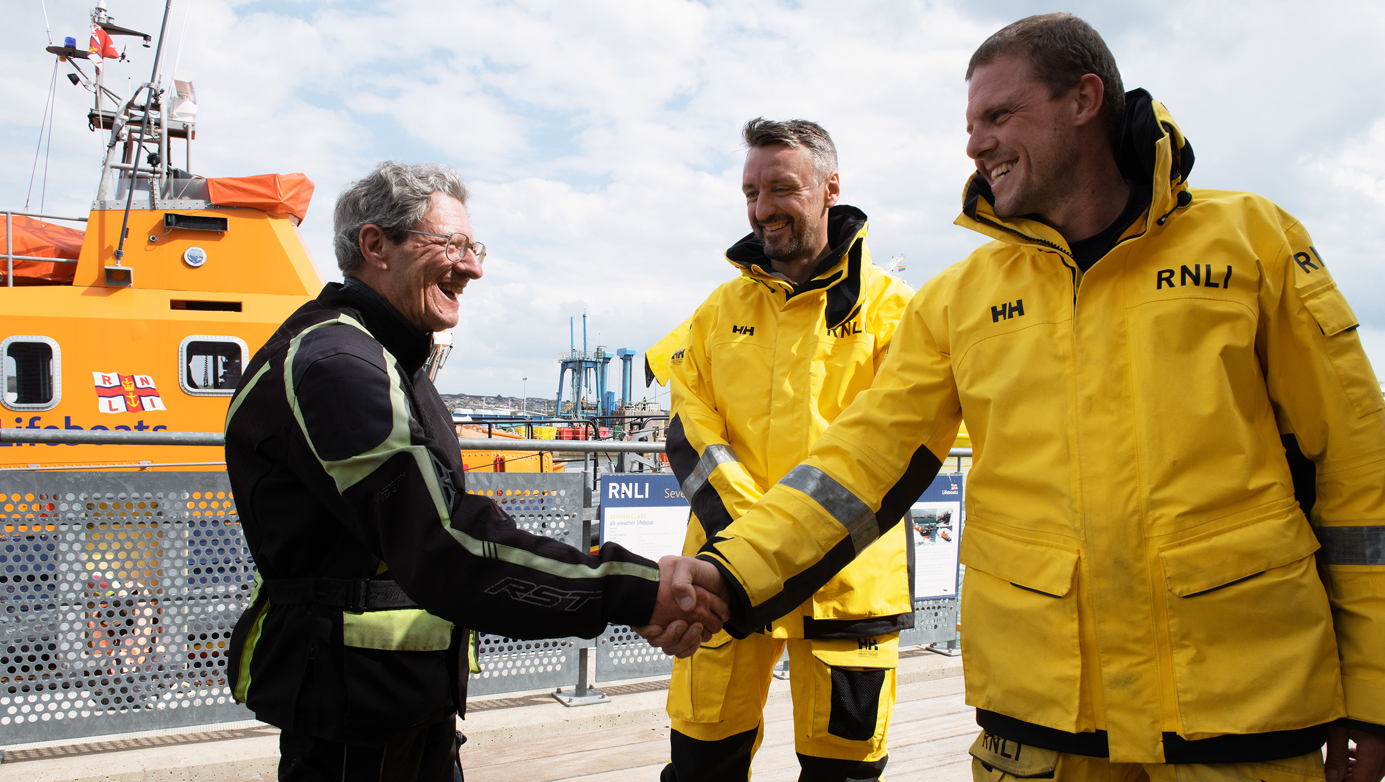 Three men are standing in front of a lifeboat. The man on the left, Dermot, is shaking hands with one of the lifeboat crew members on the right, Chris.