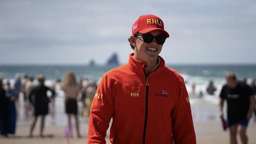Lifeguard Josh Leigh is at Perranporth Beach, wearing a red RNLI lifeguard jacket and red RNLI cap. He's looking off camera, smiling. In the background, you can see the sea and a crowd of people on the beach. 
