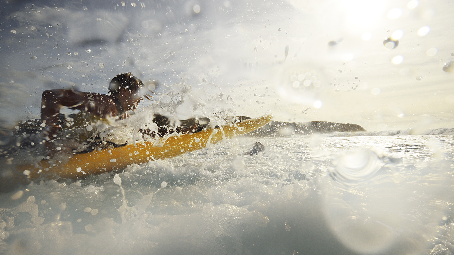 In hazy sunshine, a lifeguard plunges through the surf on the way out to sea, with surf and spray in the foreground.