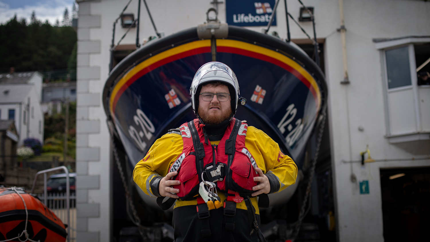 Lochlainn Leneghan, Helm at Newcastle RNLI, standing in front of the station’s all-weather lifeboat. He is wearing crew kit and is holding onto either side of the lifejacket that he is wearing. He is looking at the camera with a neutral expression.