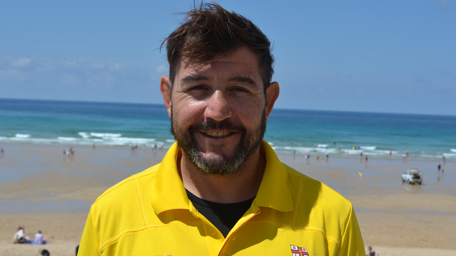 Marcelo, who has dark brown hair and a beard, smiles at the camera. He’s wearing a yellow lifeguard polo shirt. Behind him is a golden beach with turquoise waters. 