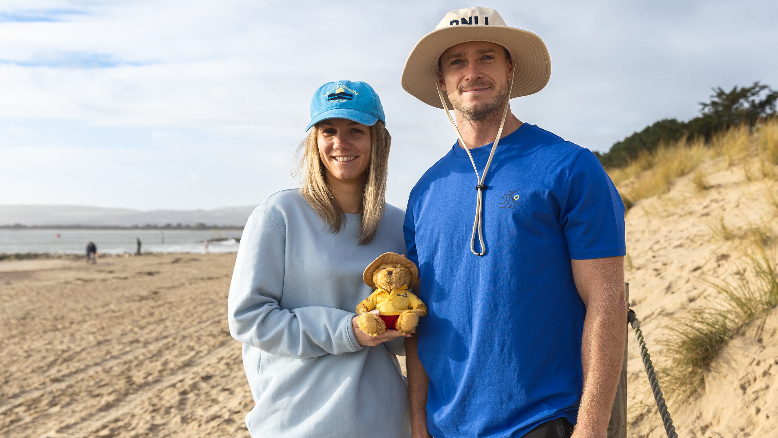 Two RNLI lifeguards, wearing pieces from RNLI Shop’s Lifeguards range, holding a lifeguard-themed teddy bear. The bear is wearing a beige boonie hat, a yellow polo top and red shorts.