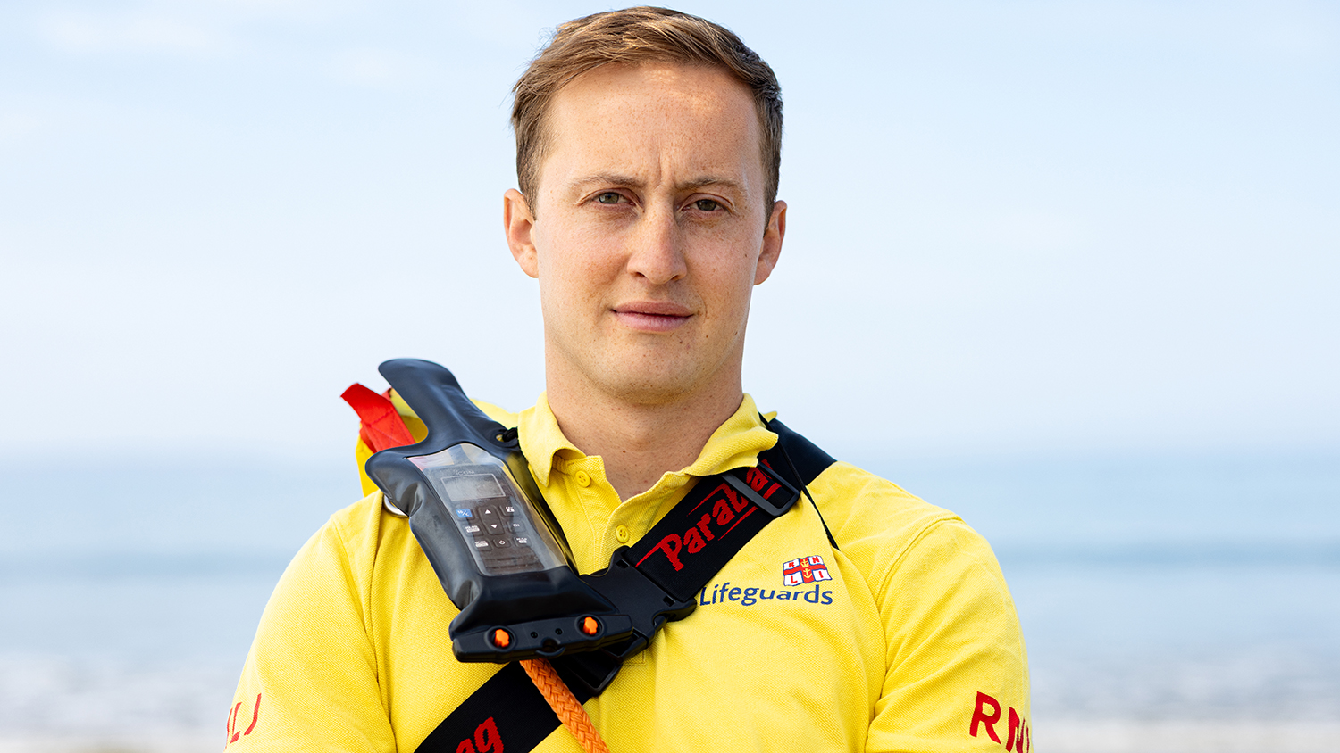 RNLI Lifeguard Matty McLeod looks towards the camera. He has a VHF radio strapped to him and a rescue tube over his shoulder.