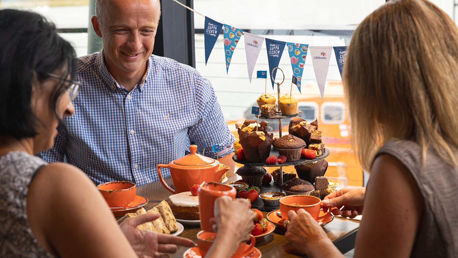 Three people are sitting around a table. Everyone is smiling and having a nice time. On the table is a cake stand filled with cakes, as well as plates covered with cakes, fruit and sweet treats.