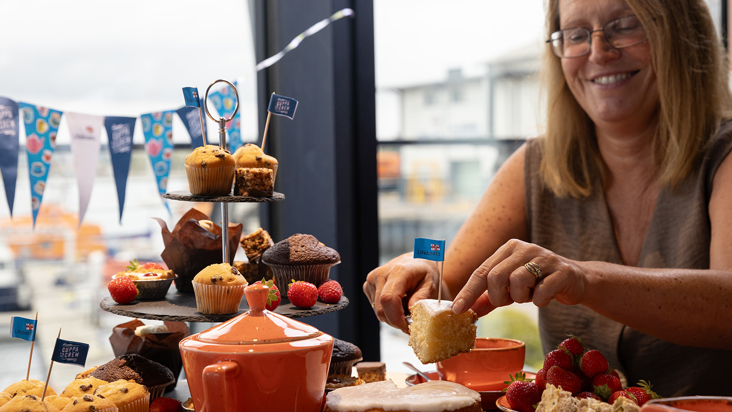 A woman is sitting at a table. A cake stand, plates of cakes and cupcakes, teacups and a teapot are all on the table. The woman, who is smiling, has cut a slice of cake and is lifting it up to place it onto a separate plate. 