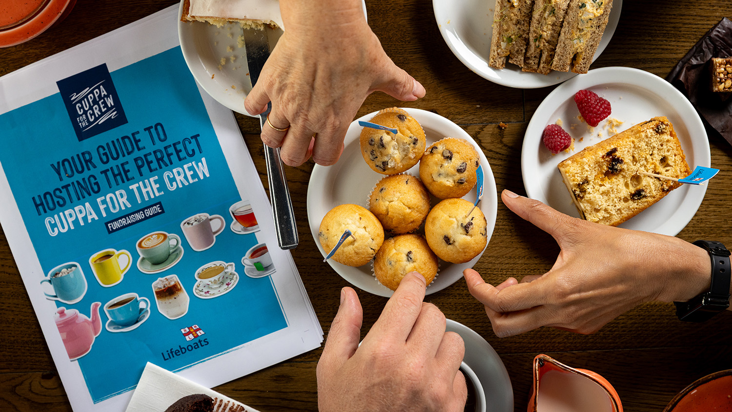 A wooden table is covered with plates of sandwiches, cakes and cupcakes. There are three hands in the shot – all three hands are reaching over to a plate of cupcakes to grab one. To the left of the plate of cupcakes, there is a Cuppa for the Crew Fundraising Guide. The photo has been taken from a top-down angle.