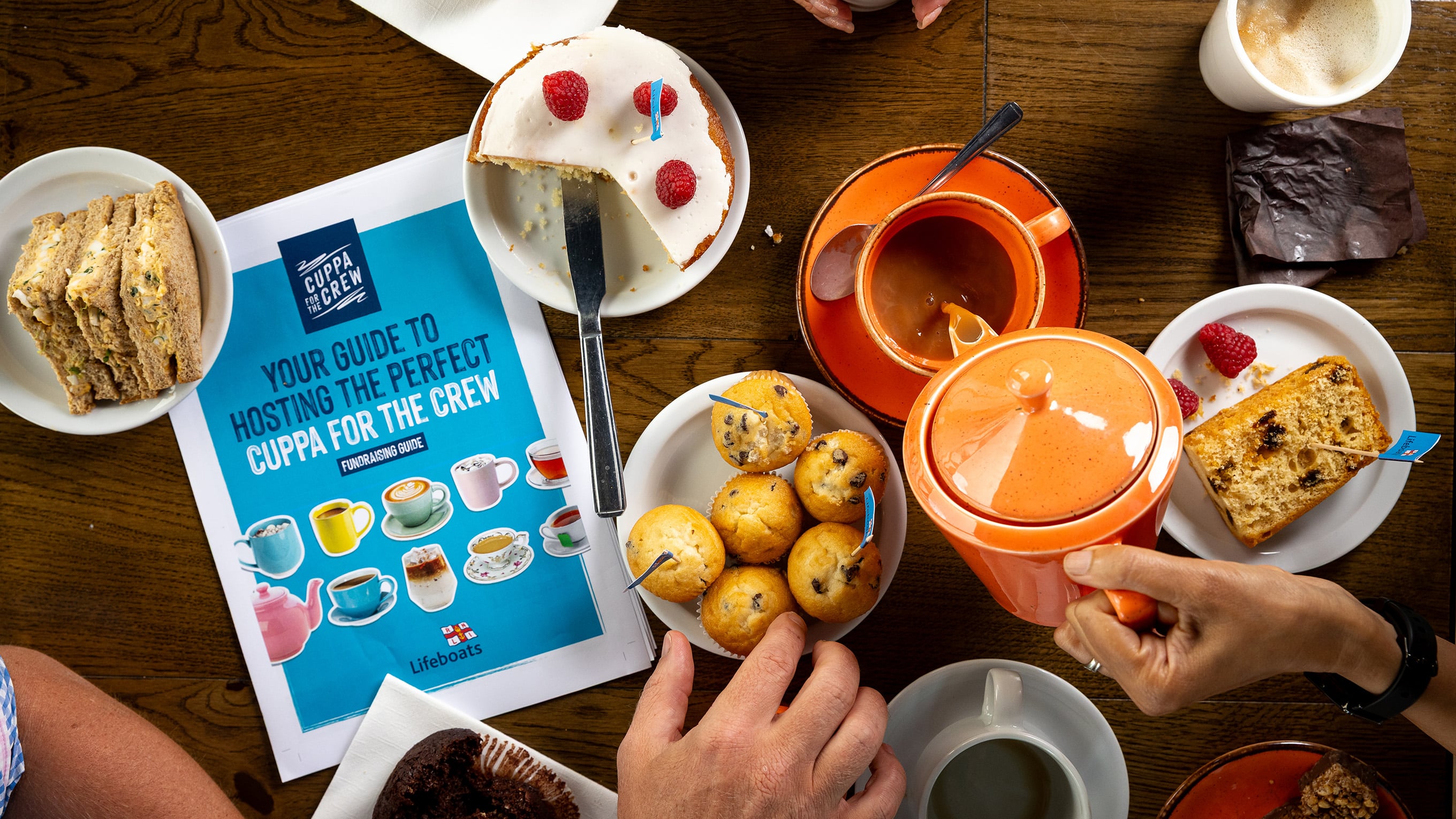 A wooden table is covered with cups of tea and coffee, as well as plates with sandwiches and cakes. There are hands in shot – one pair of hands is wrapped around a cup of tea. Another hand is reaching for a cupcake, and another hand is pouring tea from a teapot. There is also a Cuppa for the Crew Fundraising Guide on the table. The photo has been taken from a top-down angle. Photo: RNLI/Nathan Williams.