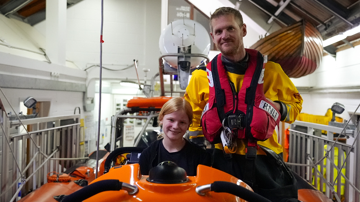 RNLI Volunteer Helm Andy Read is in Mudeford Lifeboat Station with the 11-year-old girl he rescued, on a tour of the station. The girl is sitting in the lifeboat and Andy is standing nearby. Both are smiling at the camera. 