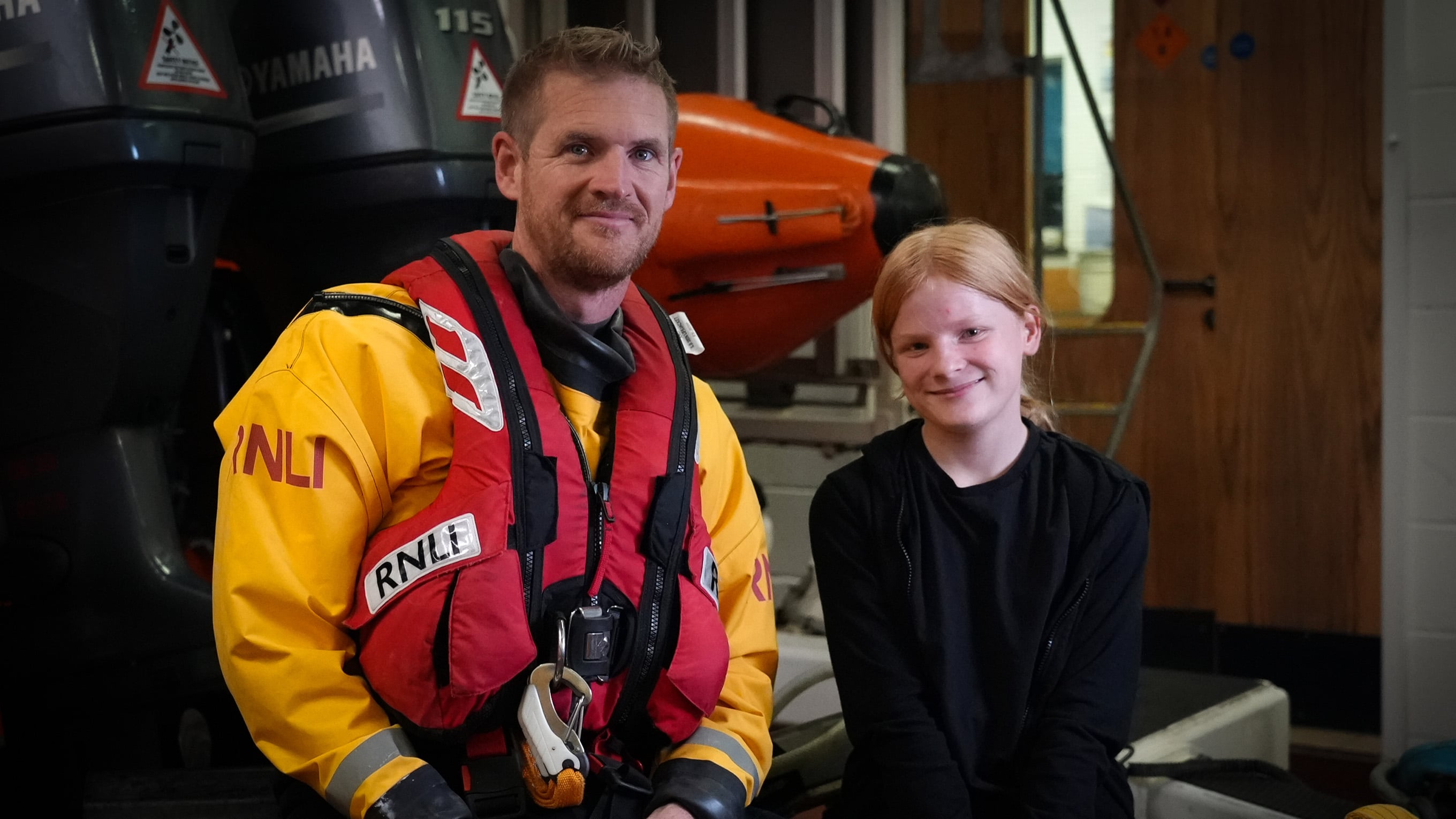Volunteer RNLI Helm Andy Read, dressed in full kit, sits in Mudeford Lifeboat Station with the 11-year-old girl he rescued. Both Andy and the girl are smiling at the camera. Photo: RNLI/Charis Jefferson. 