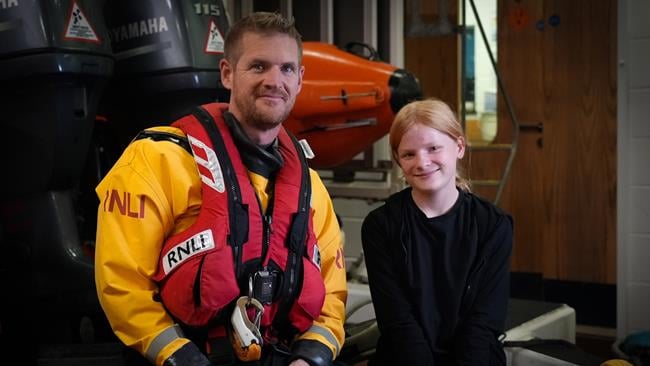 Volunteer RNLI Helm Andy Read, dressed in full kit, sits in Mudeford Lifeboat Station with the 11-year-old girl he rescued. Both Andy and the girl are smiling at the camera. Photo: RNLI/Charis Jefferson. 