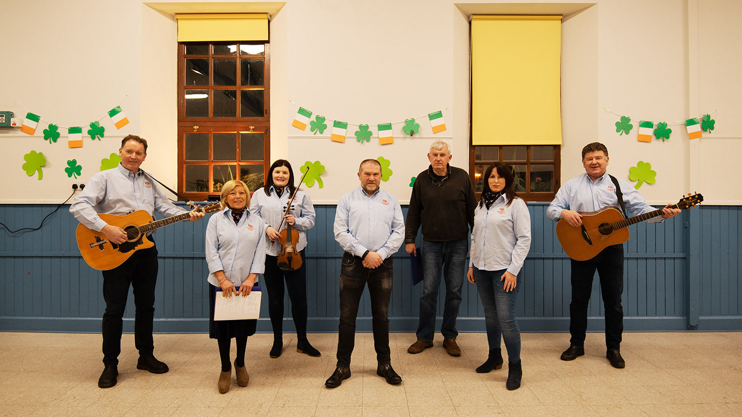Seven members of the Clogher Head choir standing together. Two are holding guitars, while one holds a violin. 