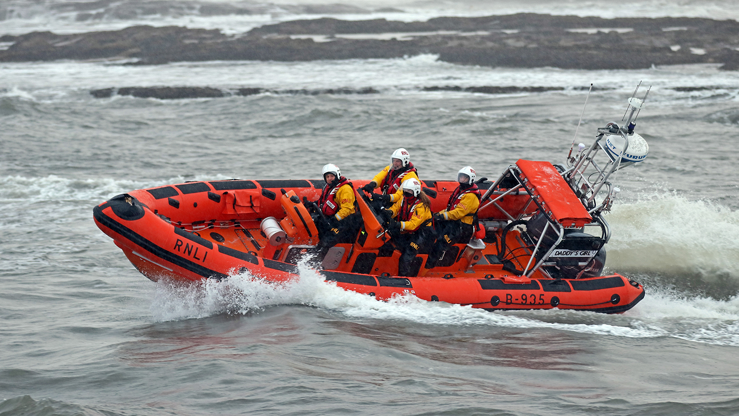 The Cullercoats crew powering through waves in their Atlantic 85 lifeboat Daddy’s Girl. The lifeboat is turning a corner in the water, causing it to tilt to the side. There are four crew members aboard the lifeboat.
