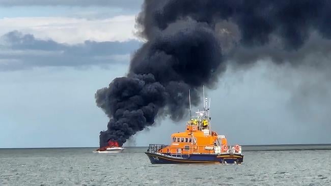 An all-weather lifeboat at sea travelling from right to left, with a white motorboat in the background giving off plumes of black smoke.
